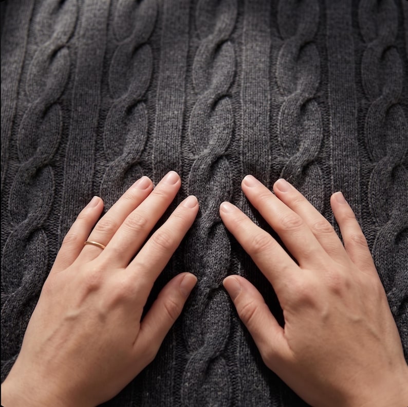May include: Close-up of a dark gray cable-knit sweater. The textured pattern of the knitwear is visible, with a series of raised, intertwined cable designs. The sweater's color is a solid, neutral gray.