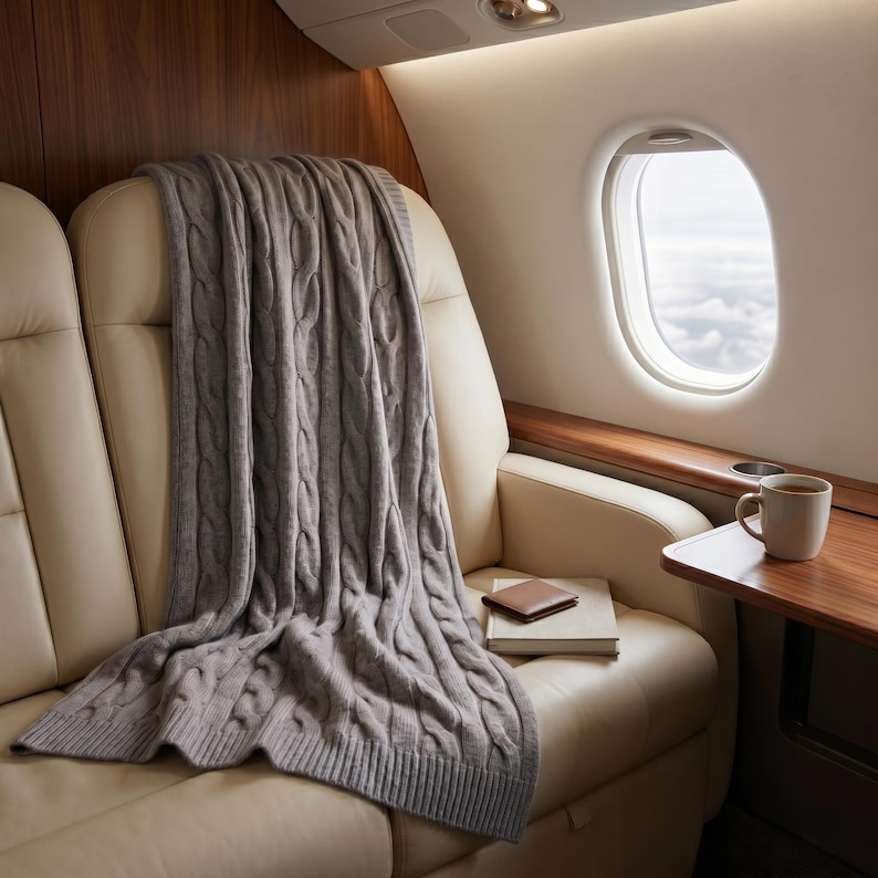 May include: A gray cable-knit throw blanket draped over a cream-colored leather seat inside an airplane. A book and a small brown leather wallet rest on the seat. A cup of coffee sits on a small wooden table next to a window with a view of the clouds.