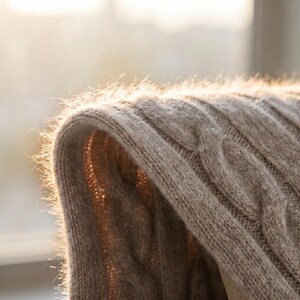 May include: Close-up of a beige cable-knit sweater. The soft, textured fabric is illuminated by sunlight, highlighting the intricate weave and the delicate fibers. The sweater's curved edge is in focus, suggesting warmth and comfort.