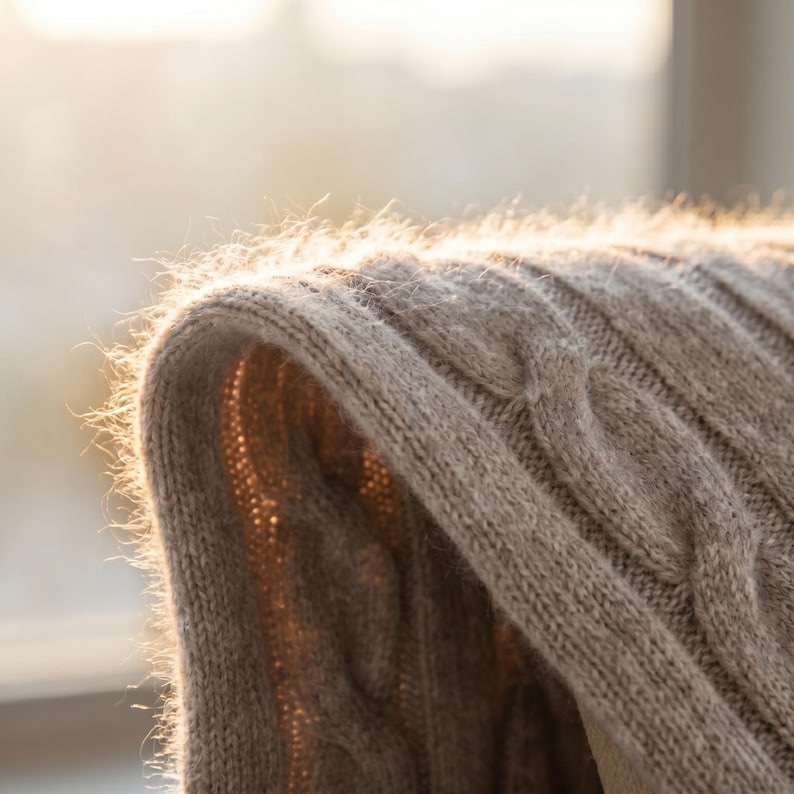 May include: Close-up of a beige cable-knit sweater. The soft, textured fabric is illuminated by sunlight, highlighting the intricate weave and the delicate fibers. The sweater's curved edge is in focus, suggesting warmth and comfort.