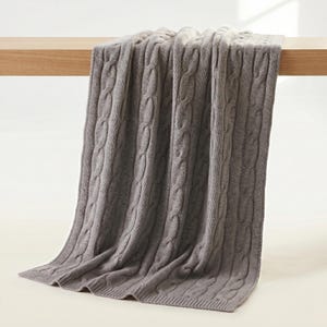 May include: A gray cable-knit throw blanket draped over a wooden surface. The blanket has a textured pattern and a soft appearance. The color is a muted gray, suitable for home decor.
