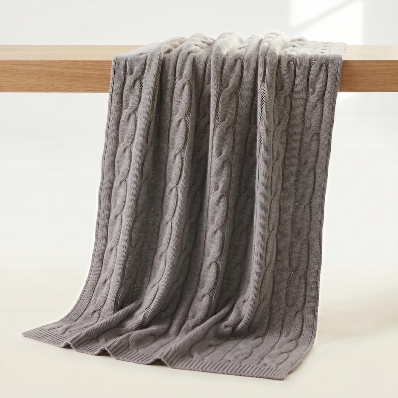 May include: A gray cable-knit throw blanket draped over a wooden surface. The blanket has a textured pattern and a soft appearance. The color is a muted gray, suitable for home decor.