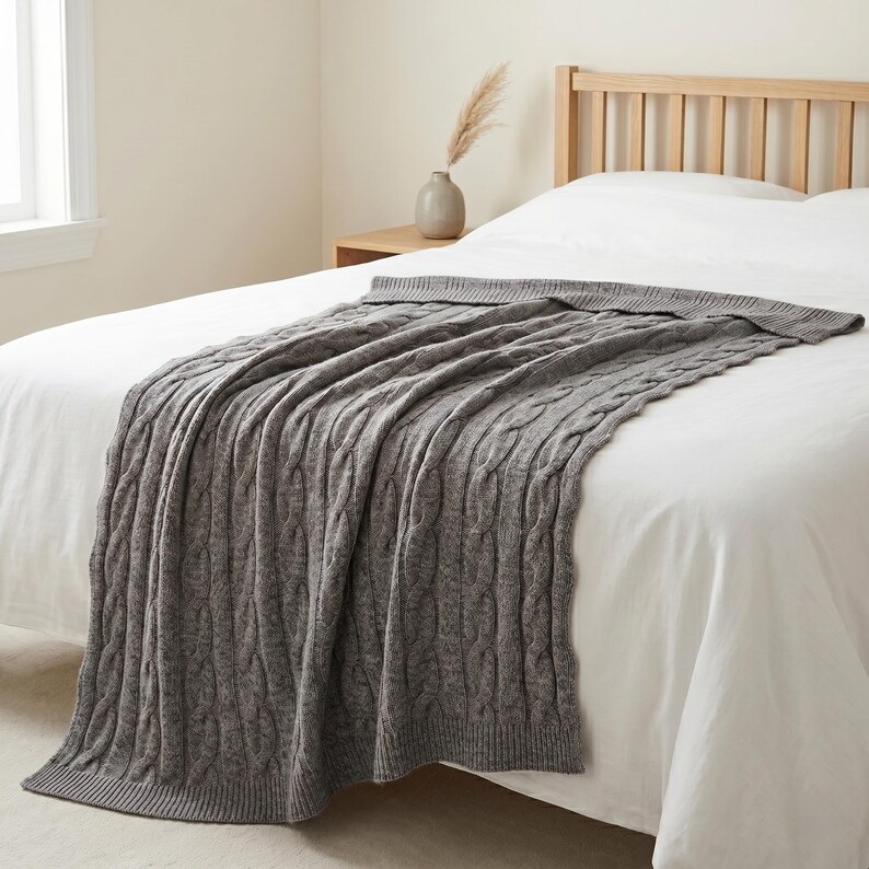 May include: A gray cable-knit throw blanket draped over a white bed. The blanket has a textured pattern and is made of a soft material. The bed is in a bedroom with a neutral color palette.
