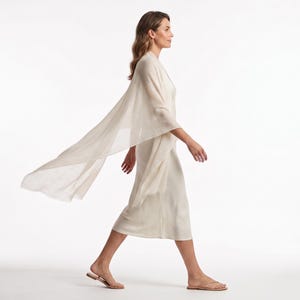 May include: A cream-colored midi-length dress with a matching lightweight, semi-sheer shawl draped over the shoulders. The dress features a simple sleeveless design with a straight hemline, paired with thin-strapped flat sandals in a neutral tone.
