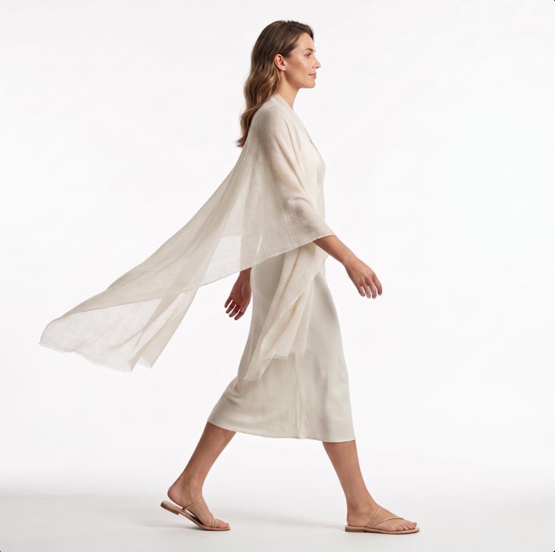 May include: A cream-colored midi-length dress with a matching lightweight, semi-sheer shawl draped over the shoulders. The dress features a simple sleeveless design with a straight hemline, paired with thin-strapped flat sandals in a neutral tone.