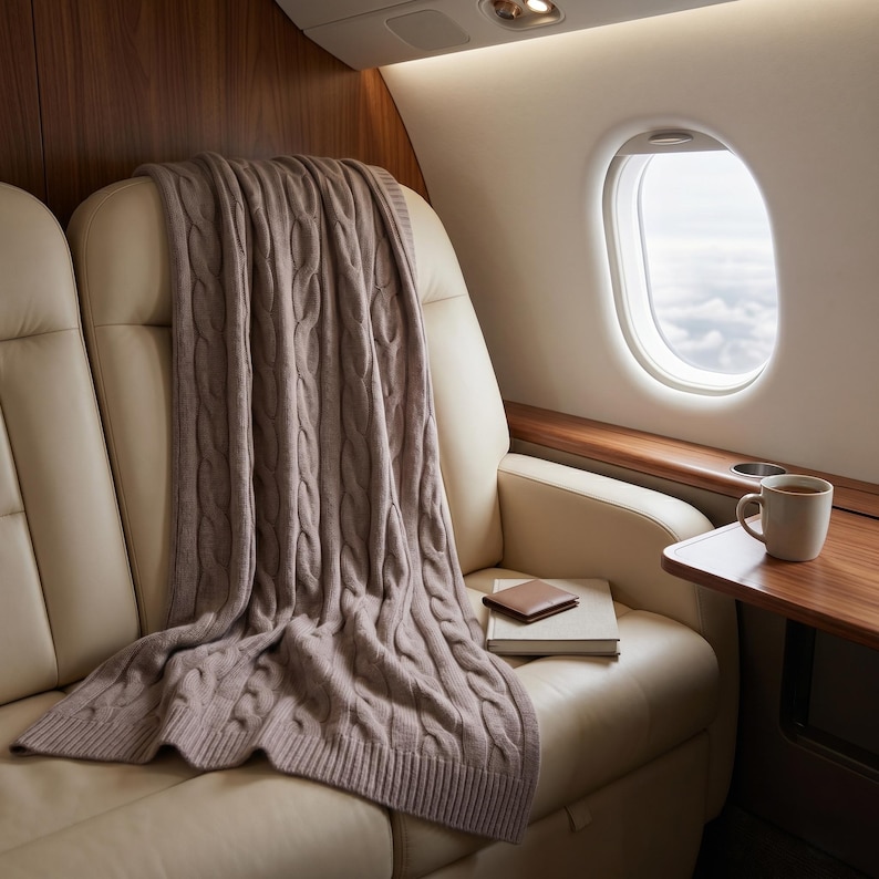 May include: A cozy, cable-knit throw blanket in a muted mauve color is draped over a cream-colored leather seat. A small brown leather wallet and a book rest on the seat. A cup of coffee sits on a small wooden table next to a window with a view of the clouds.
