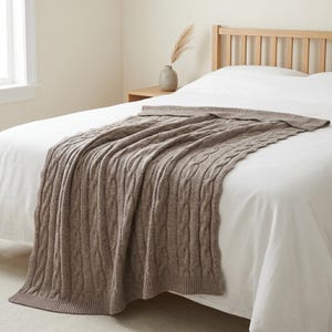 May include: A brown cable knit throw blanket draped over a bed. The blanket has a textured pattern and is made of a soft material. The bed has a white sheet and a wooden headboard. A vase with decorative grass sits on a nightstand.