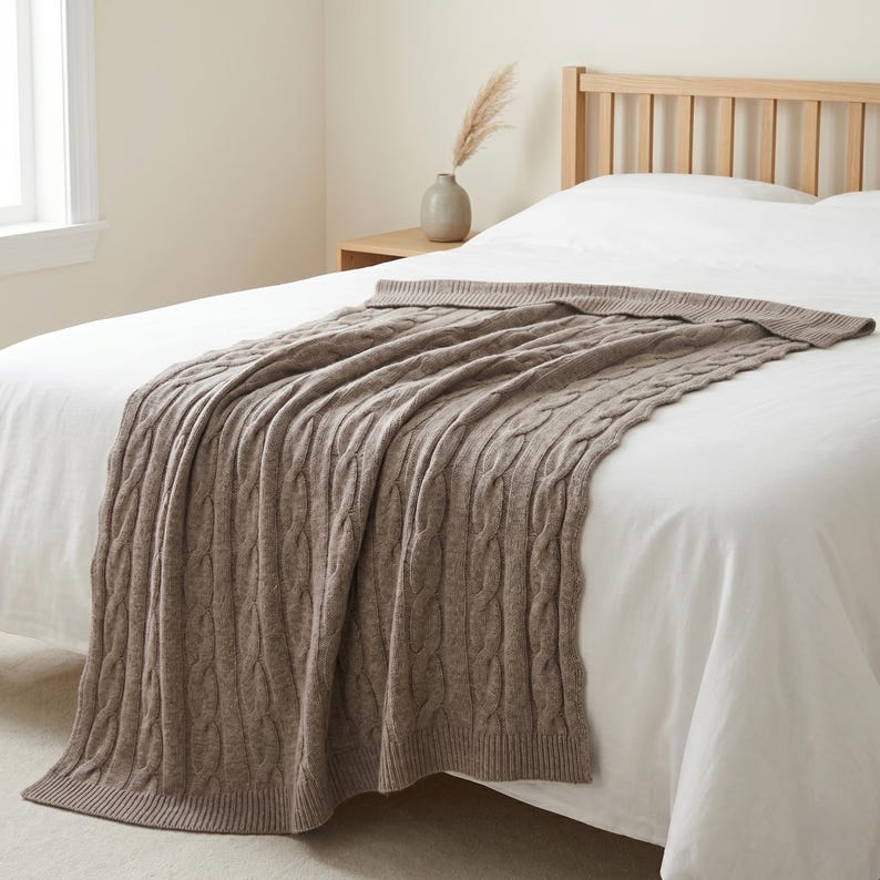 May include: A brown cable knit throw blanket draped over a bed. The blanket has a textured pattern and is made of a soft material. The bed has a white sheet and a wooden headboard. A vase with decorative grass sits on a nightstand.