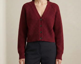 Könnte beinhalten: Eine tiefburgunderrote Langarm-Strickjacke mit gerippter Textur, V-Ausschnitt und einer Knopfleiste auf der Vorderseite mit vier passenden Knöpfen. Das Kleidungsstück wird mit einer dunkelblauen Hose kombiniert, was ein schlichtes Ensemble ergibt.