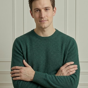 Op de afbeelding: Een donkergroene trui met ronde hals. De trui heeft lange mouwen en is gemaakt van een zacht, getextureerd materiaal. De persoon op de foto heeft de armen gekruist.