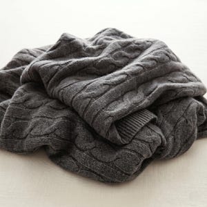 May include: A folded, charcoal gray cable-knit blanket. The blanket has a thick, textured weave with a repeating pattern of raised, braided cables. The blanket is made of a soft material and is folded over itself.
