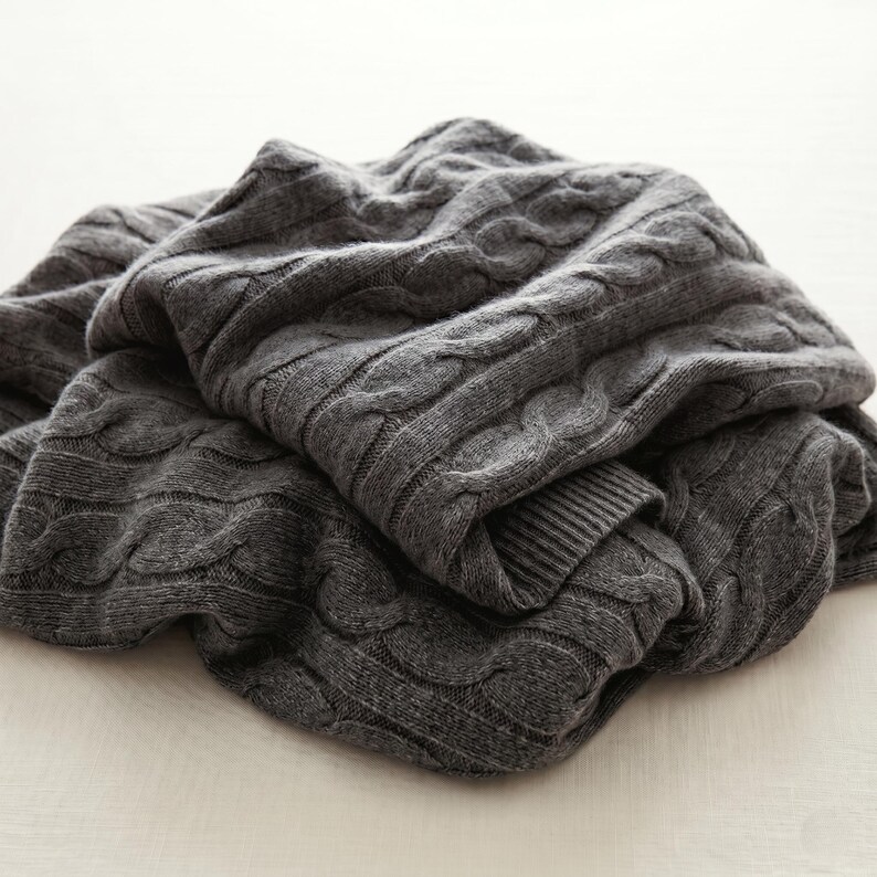 May include: A folded, charcoal gray cable-knit blanket. The blanket has a thick, textured weave with a repeating pattern of raised, braided cables. The blanket is made of a soft material and is folded over itself.