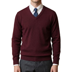 Può includere: Un maglione bordeaux con scollo a V e maniche lunghe. Il maglione è indossato sopra una camicia bianca con colletto e una cravatta blu. La persona indossa anche pantaloni grigi.