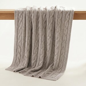 May include: A light brown cable knit blanket draped over a wooden surface. The blanket has a thick, textured weave and is made of a soft material. The blanket is folded over the edge of the wooden surface.