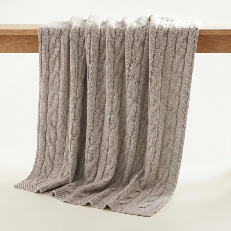 May include: A light brown cable knit blanket draped over a wooden surface. The blanket has a thick, textured weave and is made of a soft material. The blanket is folded over the edge of the wooden surface.