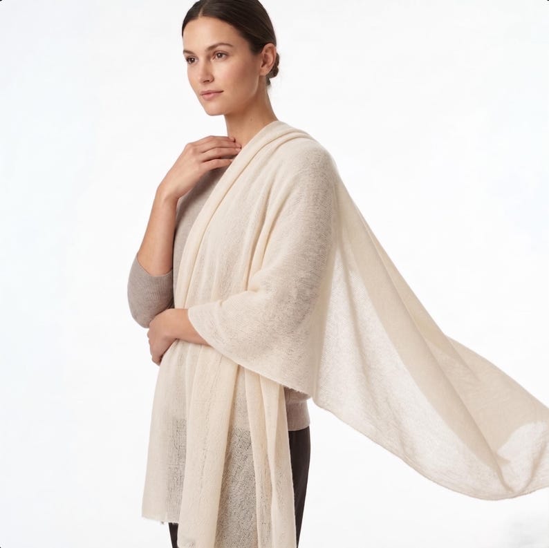 May include: A large, rectangular, cream-colored knit shawl draped over the shoulders. The fabric has a lightweight, semi-sheer texture with a fine weave, extending from the neck down to the waist and flowing outward to the side.