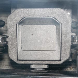 May include: Close-up of a gray, rectangular component with a textured surface and a central recessed area. The component is surrounded by metal tubes and fixtures, suggesting an automotive or industrial part.