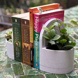 May include: A white ceramic bookend with two planters, one on each side. The bookend is holding up three books, one of which is titled "Happy Place" by Emily Henry. The bookend is sitting on a green tiled surface.