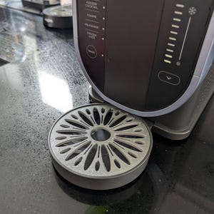 May include: A close-up of a Ninja kitchen appliance in black and silver. The control panel features settings like "Slush" and "Frozen Cocktail." A circular, grey drip tray is positioned at the base of the appliance, designed to catch drips.