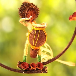 Op de afbeelding: Een grillig, handgemaakt beeldje van een persoon die trommel speelt, gemaakt van natuurlijke materialen zoals hout en touw. Het beeldje hangt aan een takachtige structuur, waardoor een decoratief mobiel ontstaat.