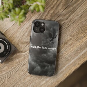 K&ouml;nnte beinhalten: Transparente Handyh&uuml;lle mit einem schwarzen und grauen Wolkenhintergrund und einer schwarzen Drachensilhouette. Der Text "well the fuck aware" ist wei&szlig; gedruckt. Die H&uuml;lle liegt auf einer Holzoberfl&auml;che.