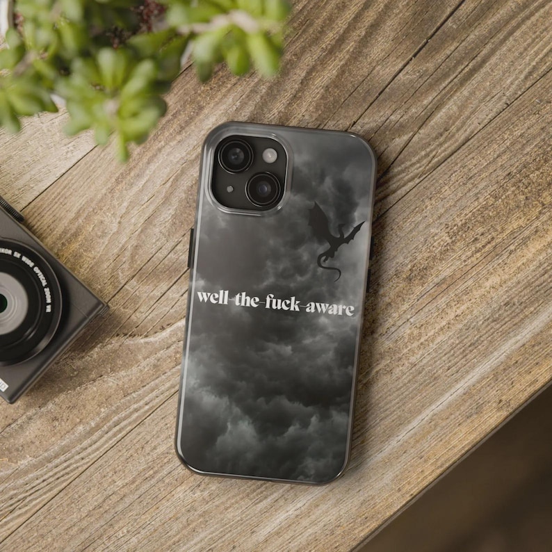 K&ouml;nnte beinhalten: Transparente Handyh&uuml;lle mit einem schwarzen und grauen Wolkenhintergrund und einer schwarzen Drachensilhouette. Der Text "well the fuck aware" ist wei&szlig; gedruckt. Die H&uuml;lle liegt auf einer Holzoberfl&auml;che.