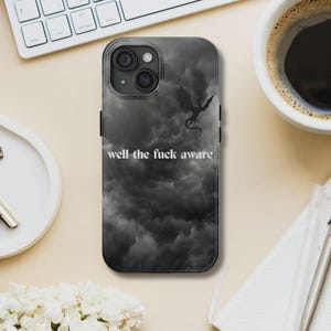 K&ouml;nnte beinhalten: Schwarze Handyh&uuml;lle mit einem dunklen, bew&ouml;lkten Himmel-Design und einer Drachensilhouette. Der Text "well the fuck aware" ist wei&szlig; gedruckt. Die Handyh&uuml;lle liegt auf einer hellen Oberfl&auml;che.