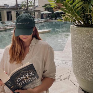 Pode incluir: Boné de beisebol verde escuro com o texto "well the f*ck aware." Uma pessoa está usando o boné e lendo um livro intitulado "Onyx Storm" de Rebecca Yarros. O fundo inclui uma piscina e palmeiras.
