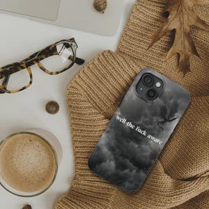 K&ouml;nnte beinhalten: Smartphone-H&uuml;lle mit dunkelgrauem Wolkenmuster und Drachensilhouette. Der Text "well the fuck aware" ist wei&szlig; aufgedruckt. Die H&uuml;lle liegt auf einem beigefarbenen Pullover.