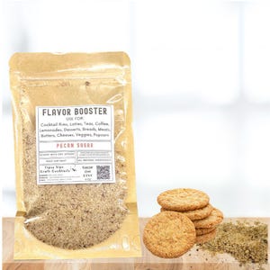 May include: A clear pouch of pecan sugar flavor booster with a white label. The label lists uses for the product, including cocktail rims, lattes, and desserts. Several round, golden-brown cookies are stacked next to the pouch.