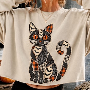 Può includere: Una felpa beige a girocollo con una grande applicazione centrale raffigurante un gatto composto da vari ritagli di tessuto. Il design del gatto presenta motivi neri, arancioni e bianchi con pipistrelli, ragnatele e quadri, rifiniti con bordi sfrangiati.