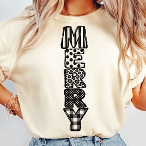 Op de afbeelding: Een crèmekleurig T-shirt met korte mouwen en het woord MERRY verticaal gedrukt in zwart-wit. Elke letter heeft een eigen patroon: M heeft verticale strepen, E stippen, R luipaardprint, R kleine stippen en Y een ruitjespatroon.
