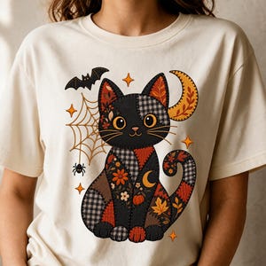 以下が含まれることがあります： クリーム色のTシャツで、チェック柄、花柄、秋の葉のパッチワーク模様でデザインされた黒猫のグラフィックが描かれています。猫の周りには、クモの巣、クモ、コウモリ、三日月、小さな四角い星が配置されています。