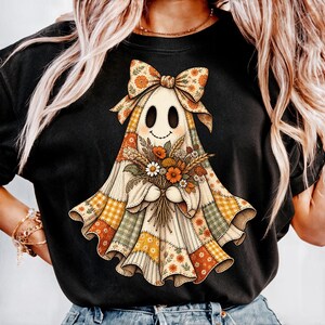 May include: A black t-shirt featuring a graphic of a ghost wearing a large bow and holding a bouquet of autumn flowers. The ghost is depicted with a patchwork-style pattern in shades of orange, yellow, and green, with a simple stitched smile.