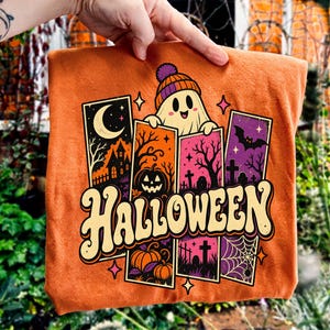 Op de afbeelding: Een oranje t-shirt met een grafische print van een lachende geest met een muts, omringd door verticale panelen met een spookhuis, een pompoen, een begraafplaats en een spinnenweb, met het woord HALLOWEEN in een groot retro lettertype.