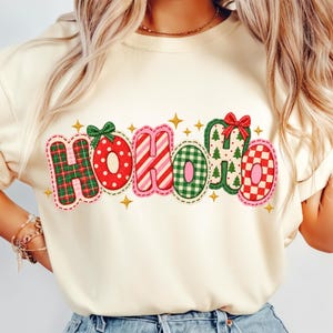 May include: A cream-colored t-shirt featuring the text HOHOHO in a festive, multi-patterned design. Each letter is outlined in pink and decorated with red and green plaid, polka dots, candy cane stripes, and Christmas tree motifs with bows.