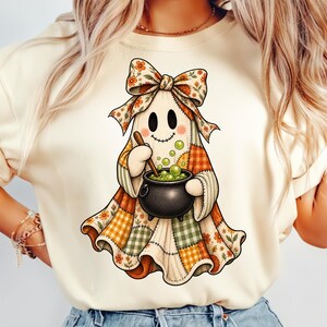 May include: A cream-colored t-shirt featuring a graphic of a ghost wearing a large floral bow and a patchwork-style robe. The ghost is holding a wooden spoon and stirring a black cauldron filled with bubbling green liquid.