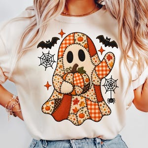 May include: A cream-colored crewneck sweatshirt featuring a central graphic of a ghost made of various autumn-themed patchwork patterns, including orange plaid, floral prints, and maple leaves. The ghost holds a pumpkin, surrounded by black bats, spiderwebs, and orange star icons.
