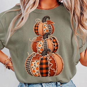 Puede incluir: Una camiseta verde oliva con un estampado gráfico de tres calabazas apiladas. Las calabazas están compuestas por varios patrones similares a telas, incluyendo cuadros escoceses naranjas y negros, rayas verticales y pequeños motivos de calabazas.