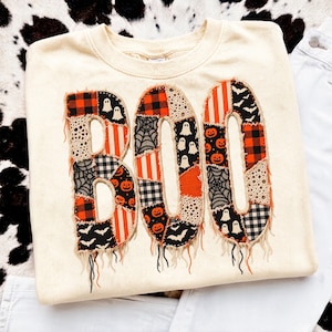 May include: A cream-coloured crewneck sweatshirt featuring the word BOO in large, appliqued letters. The letters are constructed from a patchwork of fabrics with orange, black, and white patterns including pumpkins, ghosts, bats, and plaid.
