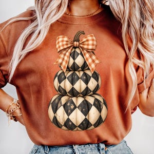 May include: A rust-colored crewneck t-shirt featuring a graphic of three stacked pumpkins. Each pumpkin has a black and cream diamond harlequin pattern. An orange and white gingham bow sits atop the stack of pumpkins on the shirt front.