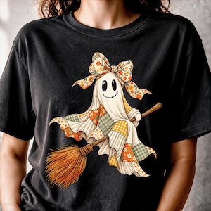 May include: A black t-shirt featuring a graphic of a white ghost wearing a floral bow and riding a wooden broomstick. The ghost is draped in a patchwork-style fabric with orange, yellow, and green checkered and floral patterns.