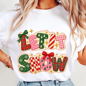 Può includere: Una t-shirt bianca a girocollo con una grafica festiva che riporta la scritta LET IT SNOW. Le lettere sono riempite con vari motivi natalizi, tra cui tartan rosso, pois rosa, alberi verdi, strisce di bastoncini di zucchero e quadri.