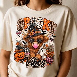 May include: A cream-colored t-shirt featuring a graphic of a Highland cow wearing a witch hat and heart-shaped sunglasses while blowing a pink bubble. The design includes pumpkins, ghosts, and the text SPOOKY VIBES in orange and black patterns.