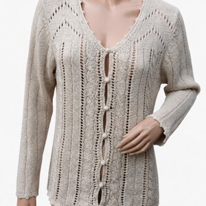 Puede incluir: Un cárdigan beige de manga larga con cuello en V, patrones de punto trenzado vertical y detalles calados. La prenda se abrocha en la parte central delantera con cinco botones pequeños, redondos y forrados en tela a juego, con borde festoneado.