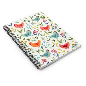 May include: Spiral-bound notebook featuring a whimsical pattern of colorful chickens, flowers, and foliage on a white background. The design includes red, orange, and blue chickens, with pink and yellow flowers. The notebook's spiral binding is black.