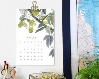 Puede incluir: Un calendario blanco con una ilustración botánica de hojas y bayas verdes. El calendario es para agosto y está colgado en una pared blanca con una grapa dorada. El calendario está al lado de un frasco de vidrio lleno de clips de papel dorados.