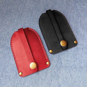 May include: Two leather key organizers, one red and one black, each featuring a rounded top, a central strap secured by a brass-colored metal snap button, and two additional brass-colored metal rivets at the bottom corners.