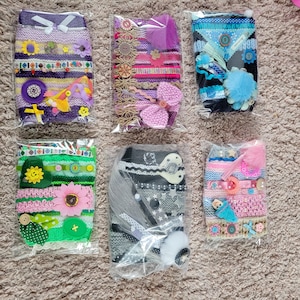 Puede incluir: Seis diademas hechas a mano en envases de plástico transparente. Las diademas presentan varios colores, incluyendo morado, verde, rosa y azul, y están adornadas con elementos decorativos como flores, lazos y botones. Cada diadema tiene adornos únicos.