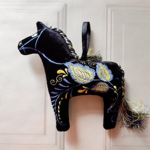 May include: A black plush horse ornament with intricate embroidery in yellow, blue, and gold. The horse has a braided mane and a tassel tail. A black ribbon loop is attached for hanging. The ornament is set against a white background.